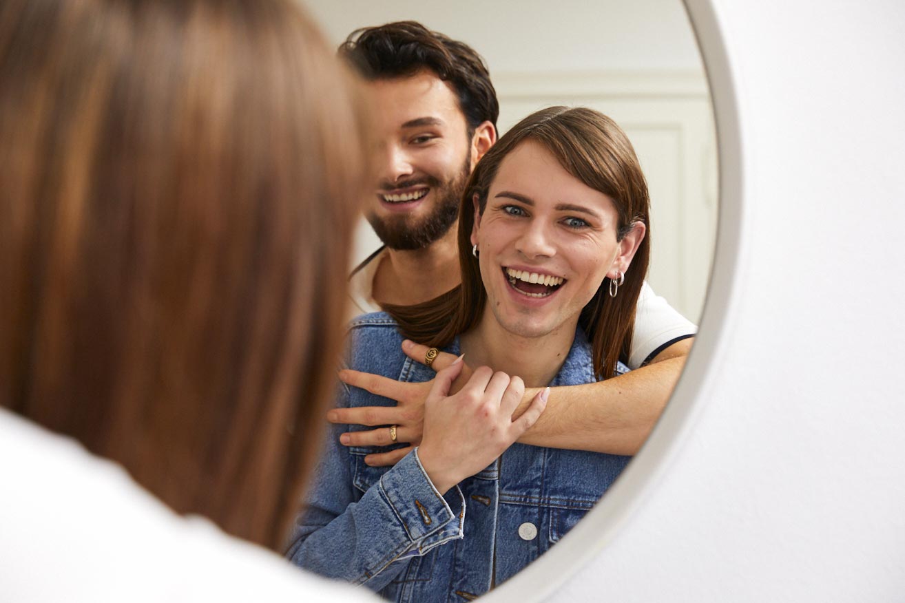 Ein queeres Pärchen umarmt sich schaut gemeinsam glücklich in einen Spiegel.