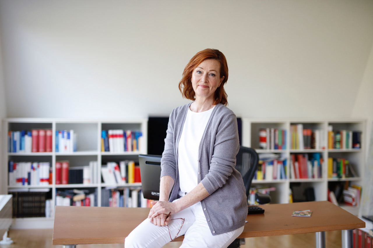 Eine Ärztin steht angelehnt an ihrem Schreibtisch und lächelt.