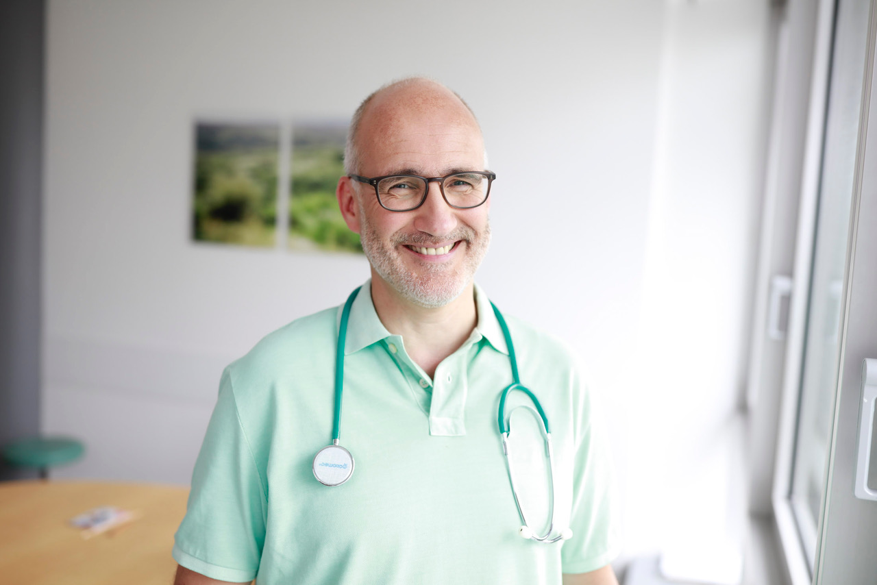 Ein Arzt steht in seinem Büro und lächelt freundlich in die Kamera.