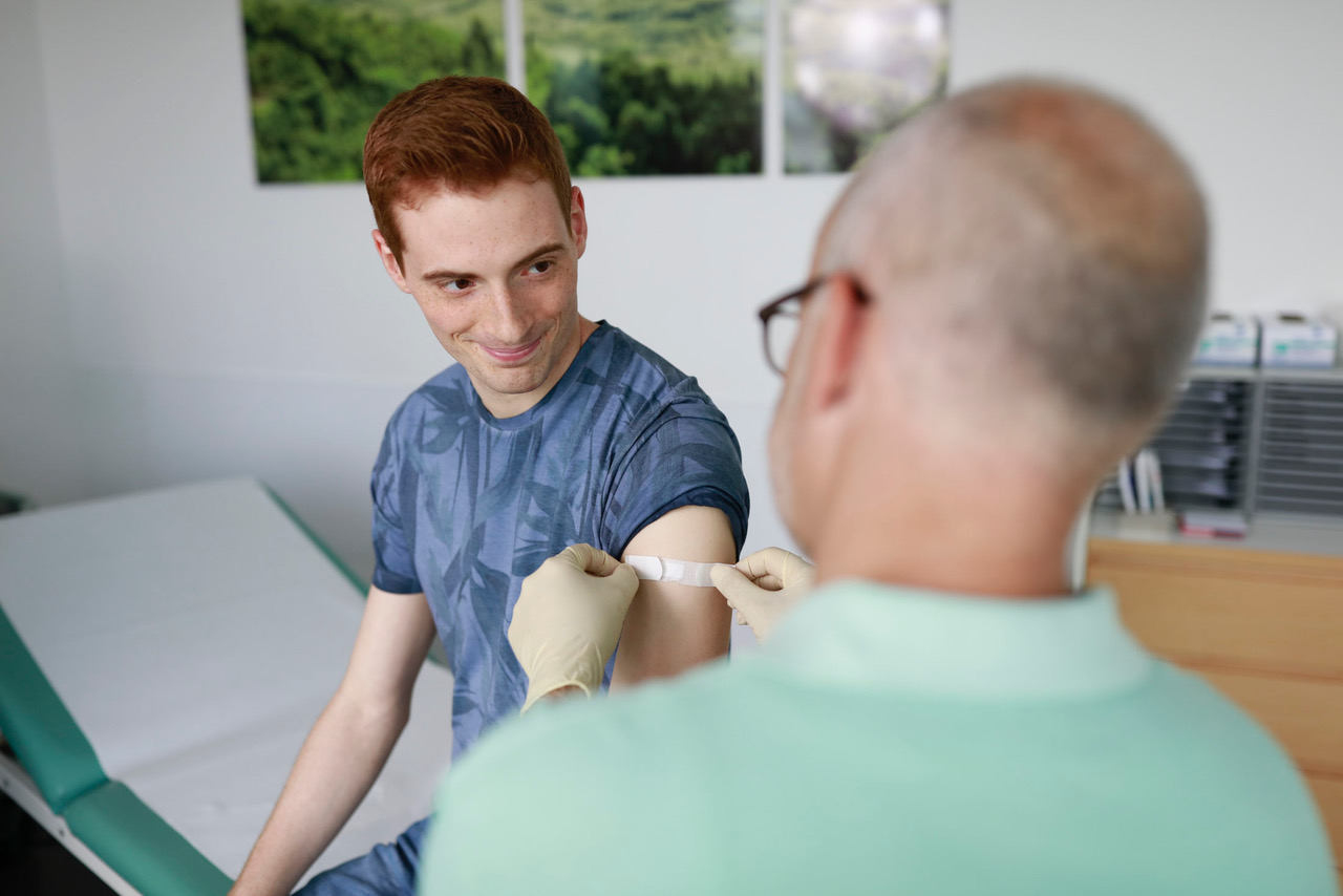 Das Bild zeigt einen jungen Mann beim Arzt, der ihm gerade ein Pflaster auf den Oberarm klebt.