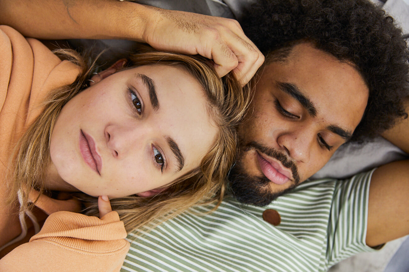 Eine junge Frau liegt in den Armes eines jungen Mannes und blickt verträumt nach oben.