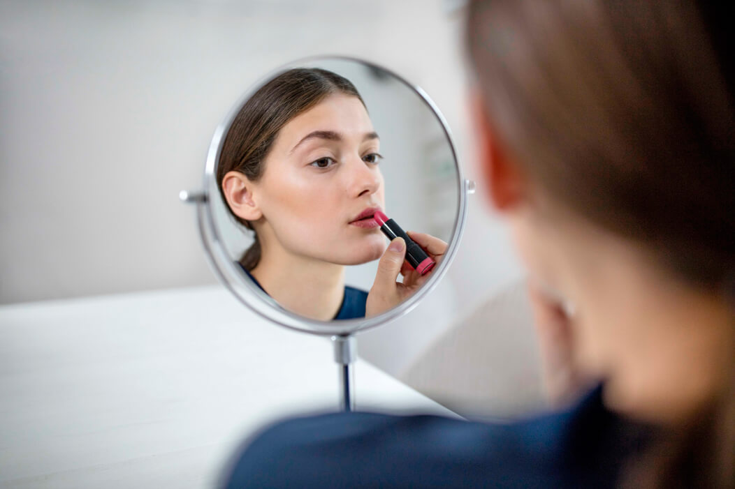 Eine junge Person schaut in einen Spiegel und trägt Lippenstift auf.