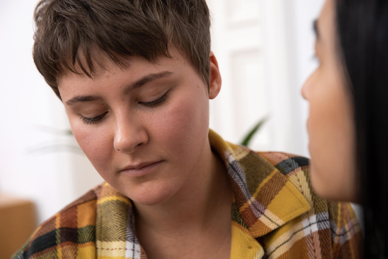 Eine junge Person schaut nachdenklich nach unten. Ihre Partnerin ist im Anschnitt zu sehen.