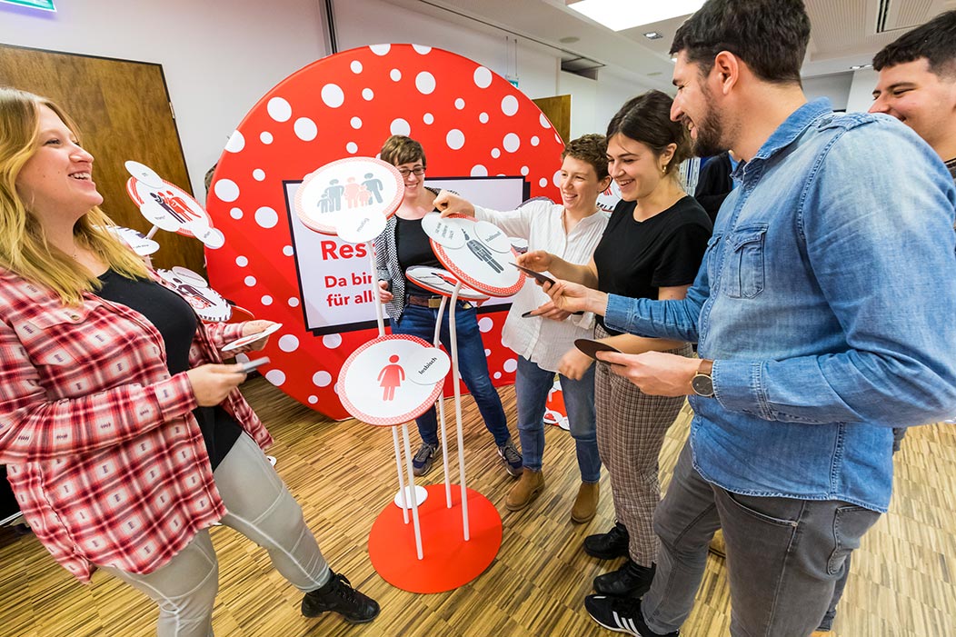 Eine Gruppe Moderierender steht lachend um einen Aufsteller der Station "Vielfalt und Respekt". Sie hängen Karten mit Begriffen zu Illustrationen an dem Aufsteller.
