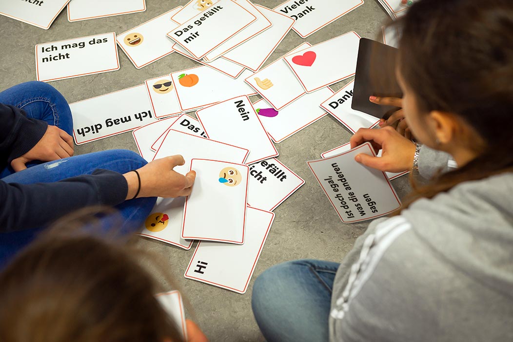 Auf dem Boden liegen Karten mit Emojis und Textbausteinen, zum Beispiel "Das gefällt mir nicht". Drumherum sitzen Schülerinnen. Manche haben eine der Karten in der Hand.