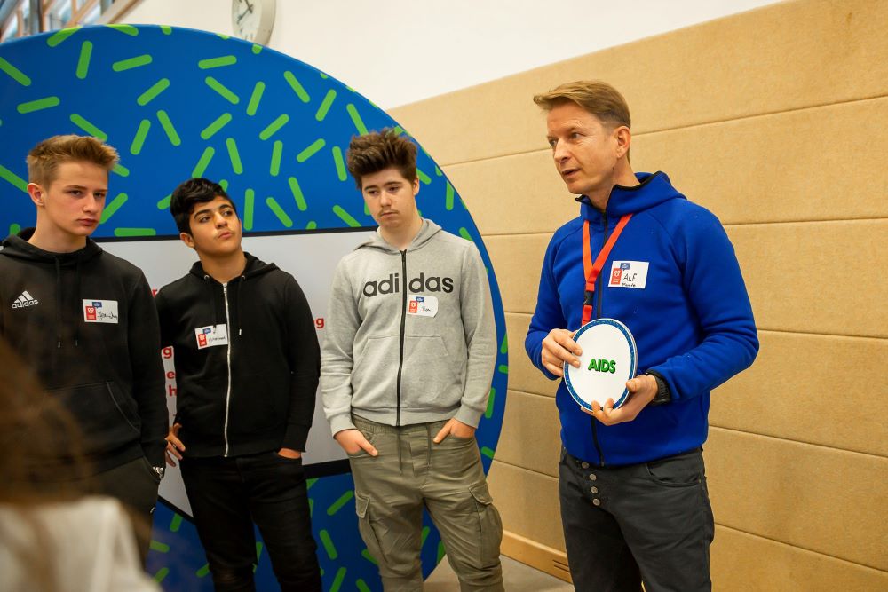 Ein Moderator hat eine runde Karte mit der Aufschrift »AIDS« in der Hand. Drei Schüler stehen neben ihm.