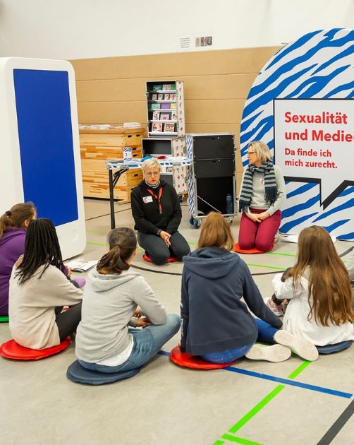Fünf Schülerinnen sitzen im Halbkreis um eine Moderierende und eine Lehrkraft. Auf einem großen Aufsteller dabene steht "Sexualität und Medien. Da finde ich mich zurecht."