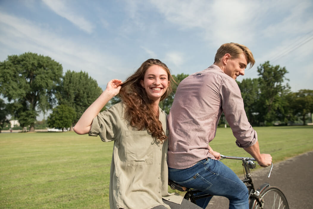 Junger Mann radelt mit seiner Partnerin auf dem Gepäckträger durch einen Park, beide lachen fröhlich.