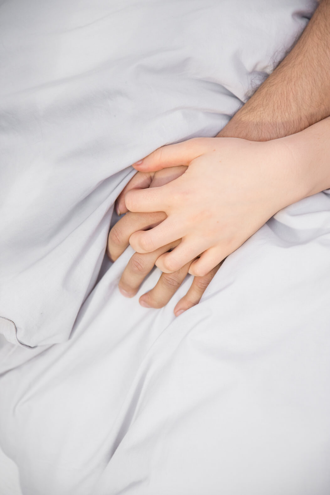 Im Anschnitt: Hände einer Frau und eines Mannes liegen auf einer Bettdecke und greifen ineinander.