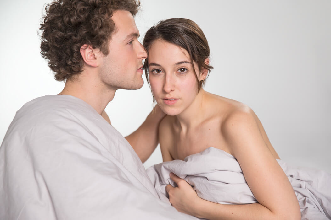 Heterosexuelles Paar unbekleidet unter einer Bettdecke mit ernstem, betroffenem Blick.