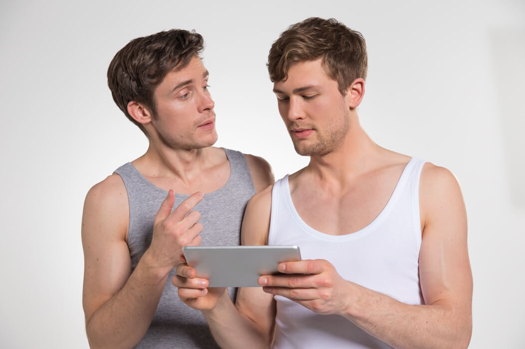 Schwules Paar in leichter Bekleidung schaut gemeinsam auf ein Tablet, einer der Männer erklärt etwas.