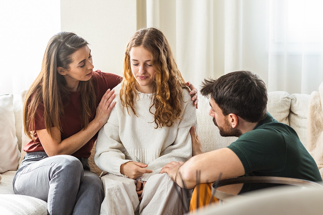 Das Bild zeigt ein junges Mädchen auf dem Sofa sitzend, das nach nachdenklich nach vorne schaut. Rechts von ihr kniet ihr Vater neben dem Sofa und sieht sie an, links von ihr sitzt ihre Mutter, die die Tocher an der der Schulter berührt.